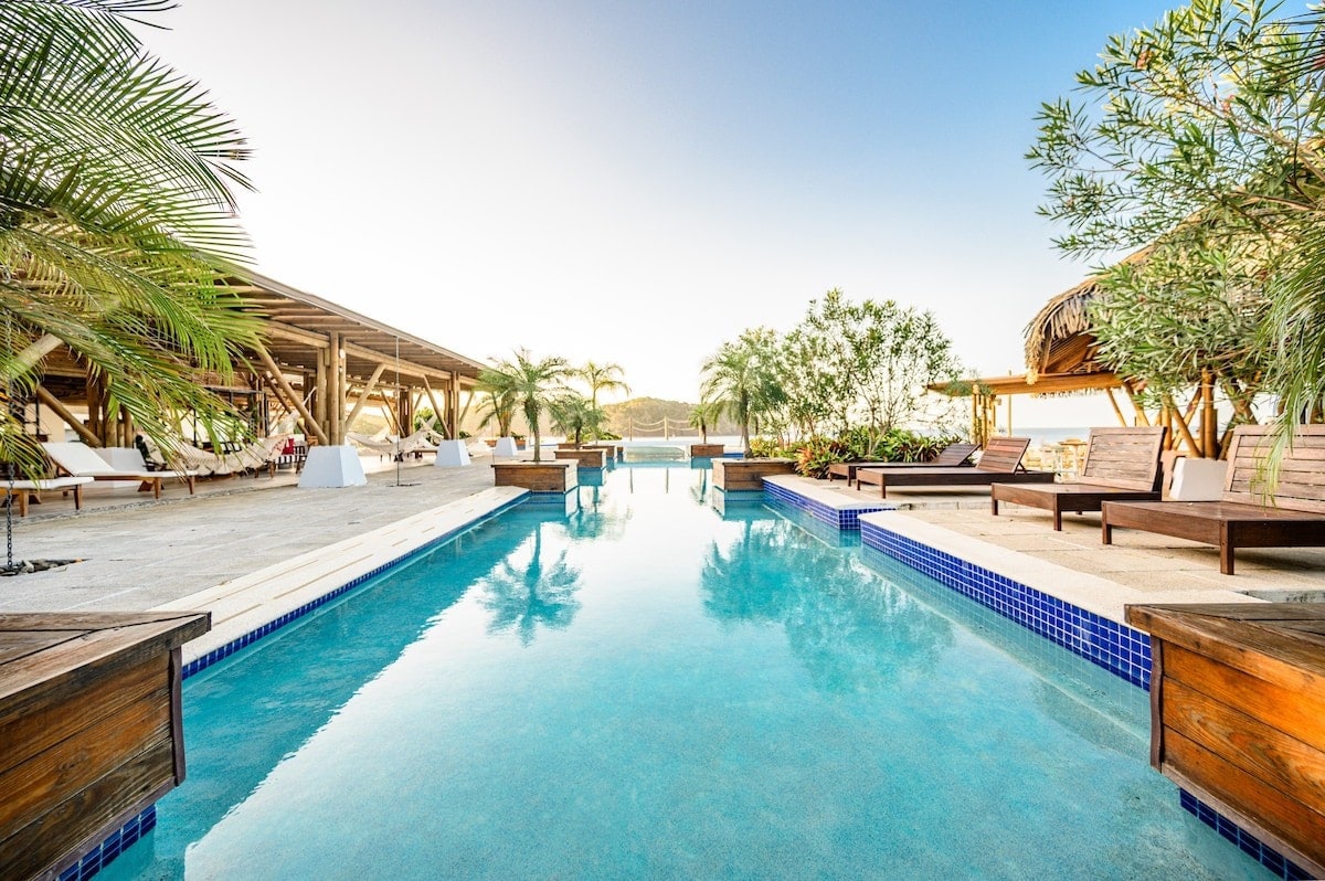A serene infinity pool stretches toward the horizon, surrounded by lush palm trees and comfortable loungers. The pool's clear water reflects the open sky, inviting relaxation. A wooden structure offers shaded seating areas, creating a tranquil environment for guests to unwind.