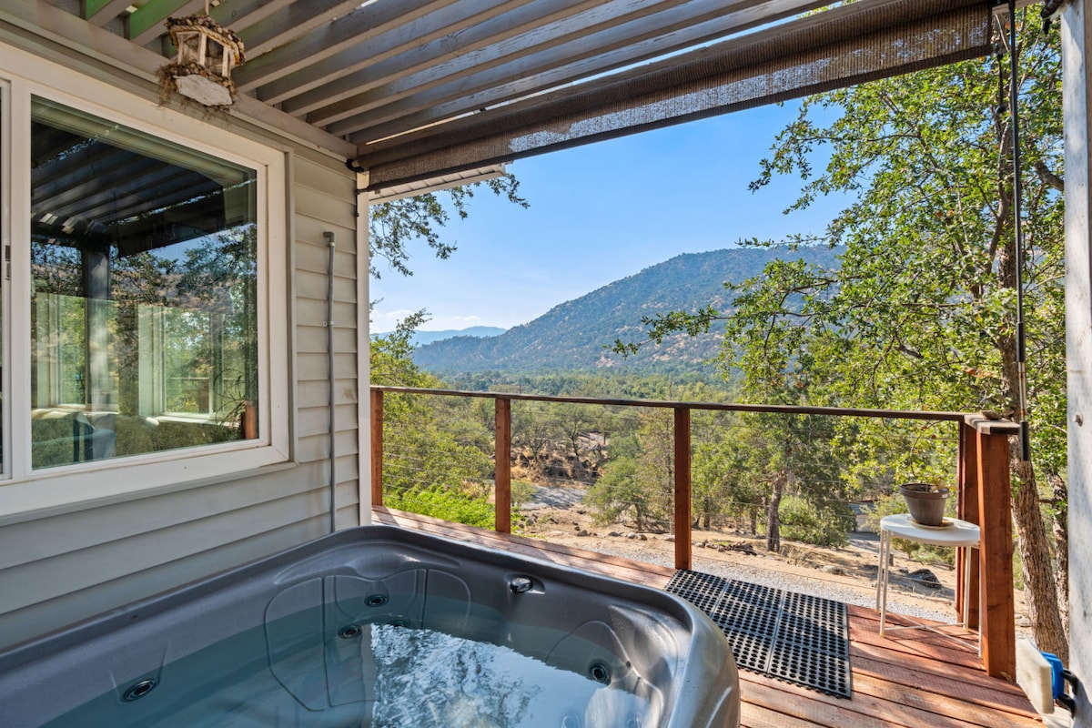 A hot tub is positioned on a wooden deck, offering expansive views of the surrounding mountains and tree-filled landscape. Large windows provide natural light, while a small table with a potted plant sits nearby, contributing to a serene outdoor ambiance.