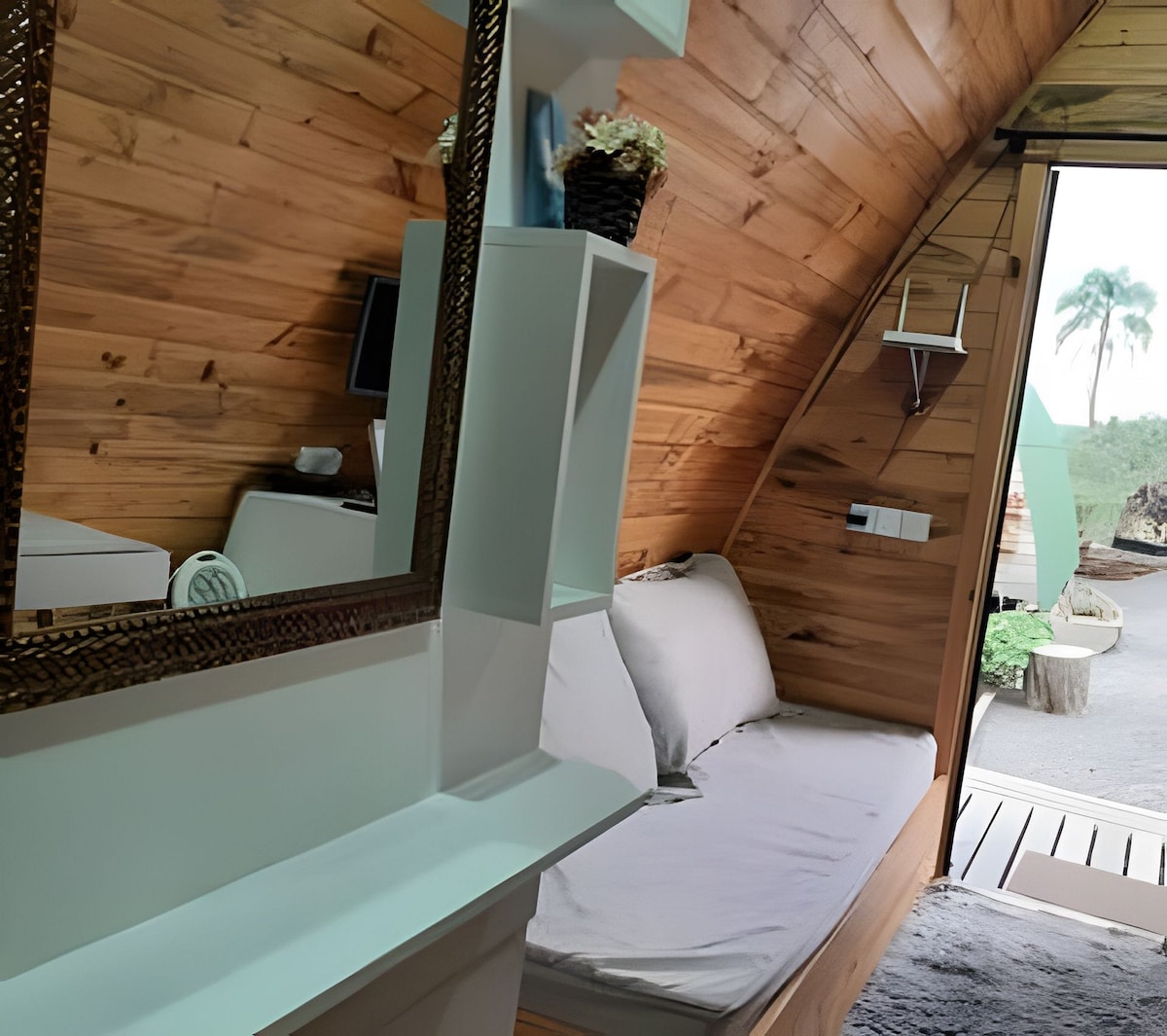 A modern interior is showcased with wooden walls and a cozy seating area, featuring a minimalistic design. A mirror adds depth to the space, while natural light enters through an opening, illuminating the room. A television is mounted on the wall for entertainment.