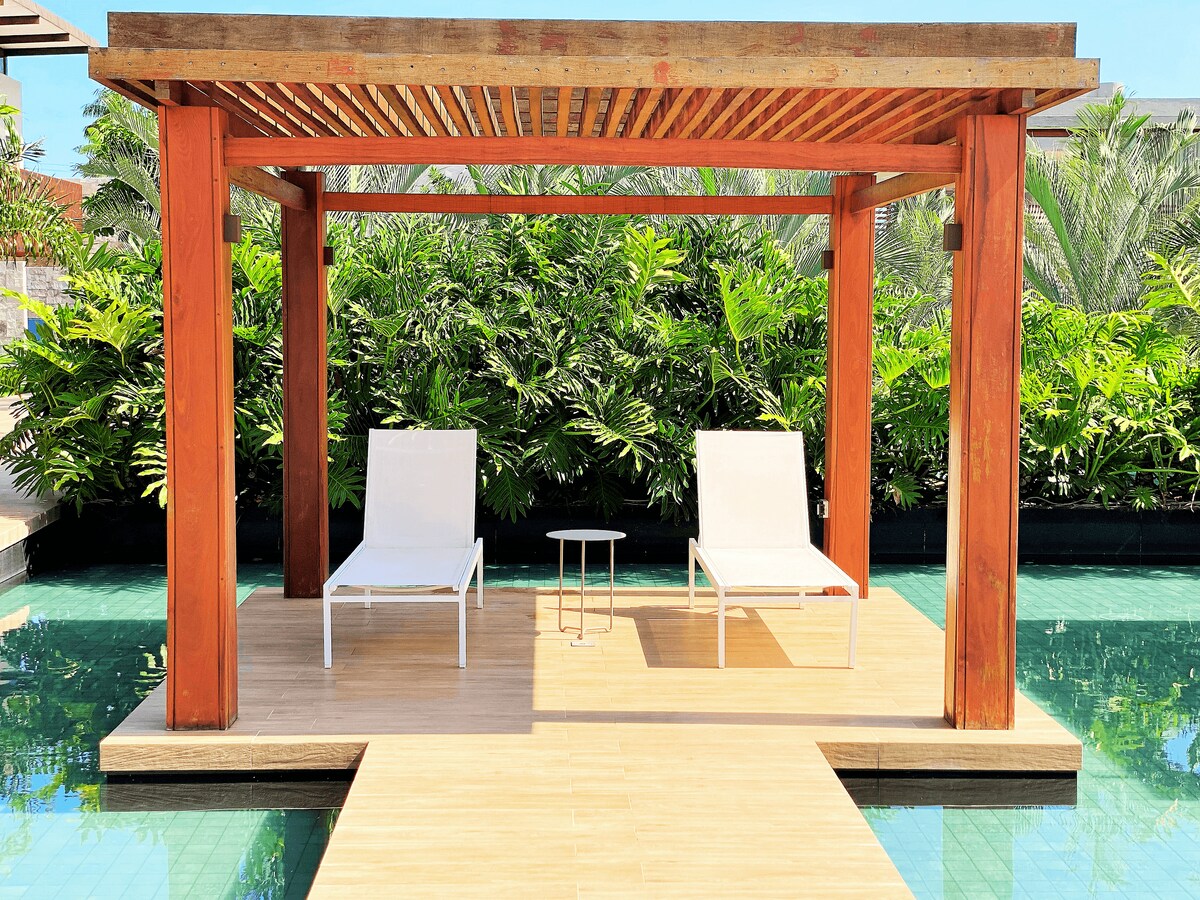 A wooden pergola frames a sunlit area adjacent to a tranquil pool, featuring two white lounge chairs and a small side table. Lush greenery surrounds the space, contributing to a serene atmosphere for relaxation.
