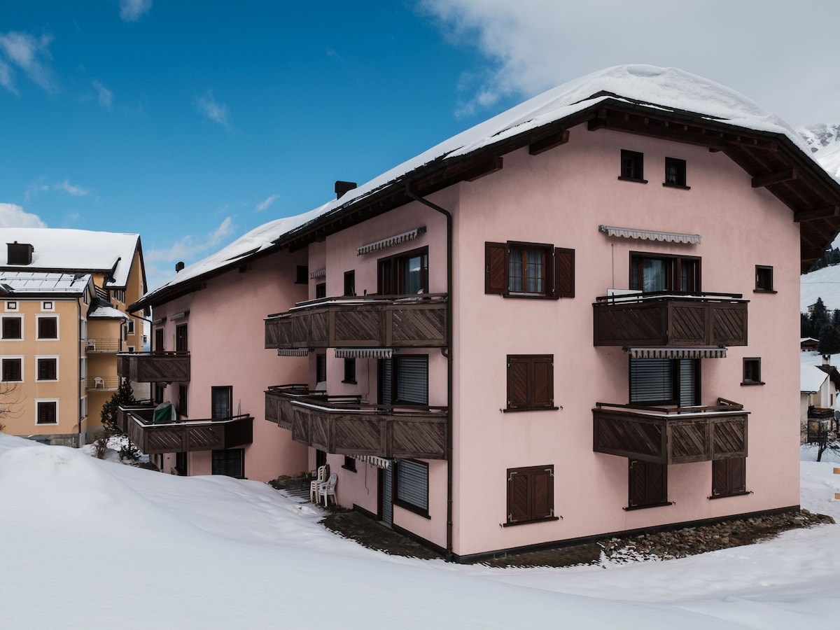 A large, three-story apartment building with a pink exterior, featuring multiple balconies with wooden railings. Snow covers the ground around the building, and a clear blue sky is visible above, highlighting the mountain backdrop.