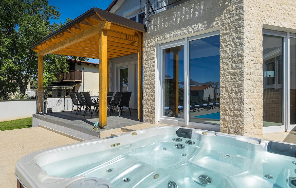 An outdoor area features a modern hot tub adjacent to a covered terrace with a dining table and chairs. Large glass doors showcase the villa's interior, enhancing the connection between indoor and outdoor spaces. Lush greenery can be seen in the background.