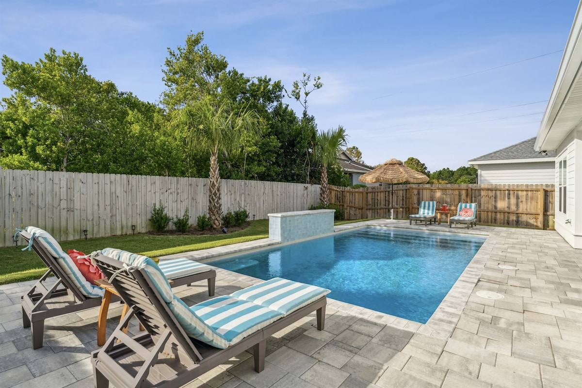 A spacious backyard features a heated pool surrounded by lounging chairs with striped cushions. Lush greenery and palm trees provide privacy, while thatched umbrellas add a tropical feel, making the area ideal for relaxation and outdoor gatherings.