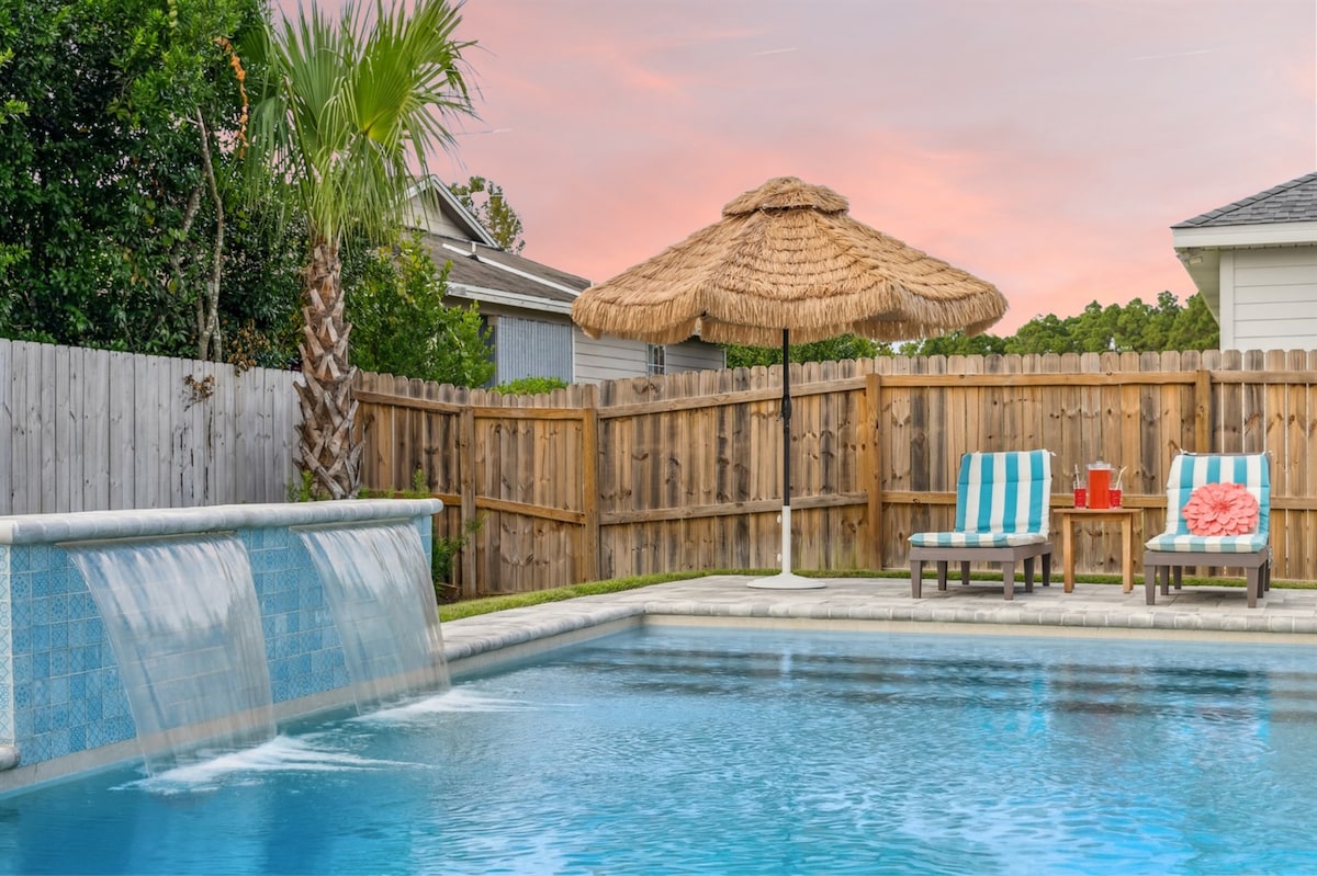 A spacious pool area is showcased with a heated pool featuring water features and a straw-covered umbrella. Two lounge chairs are positioned nearby on a stone patio, accompanied by a small table holding a drink and a vibrant decorative pillow.