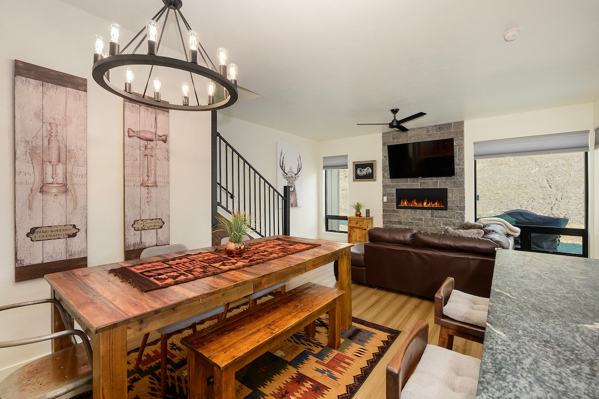 An inviting dining area features a large wooden table surrounded by chairs, complemented by a rustic chandelier overhead. A cozy living room is visible, equipped with a sectional sofa and a gas fireplace beneath a mounted television, with large windows allowing natural light.