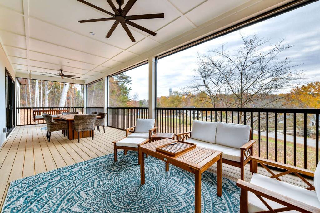 A spacious screened deck is featured with comfortable seating, including light-colored sofas and wooden armchairs. A patterned area rug adds color underfoot. Large ceiling fans and natural light enhance the relaxing atmosphere while views of the surrounding trees are visible through the screens.