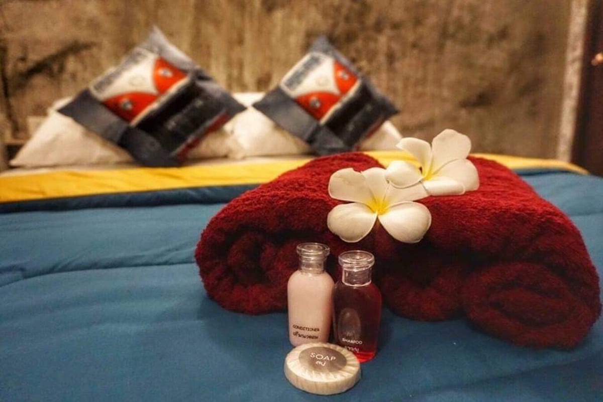 A neatly arranged bed is seen in the image, featuring dark blue and yellow bedding. Red rolled towels lie at the foot of the bed, accompanied by small bottles of shampoo, conditioner, and soap. Freshly placed flowers add a touch of natural beauty to the scene.
