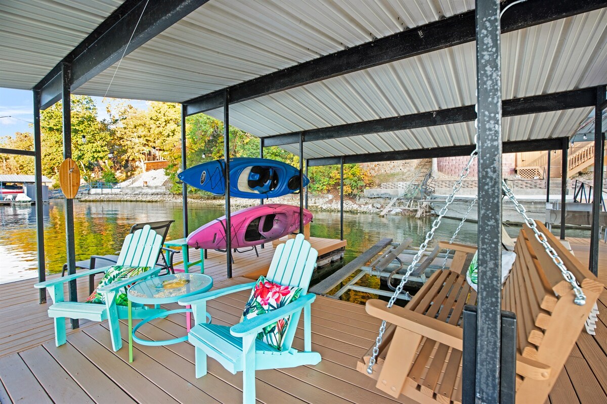 A covered dock area features colorful kayaks suspended overhead, complementing the serene lake view. Seating arrangements include two turquoise chairs and a wooden swing, enhancing comfort for relaxation. A round table is adorned with a decorative centerpiece, inviting casual gatherings in this lakeside retreat.
