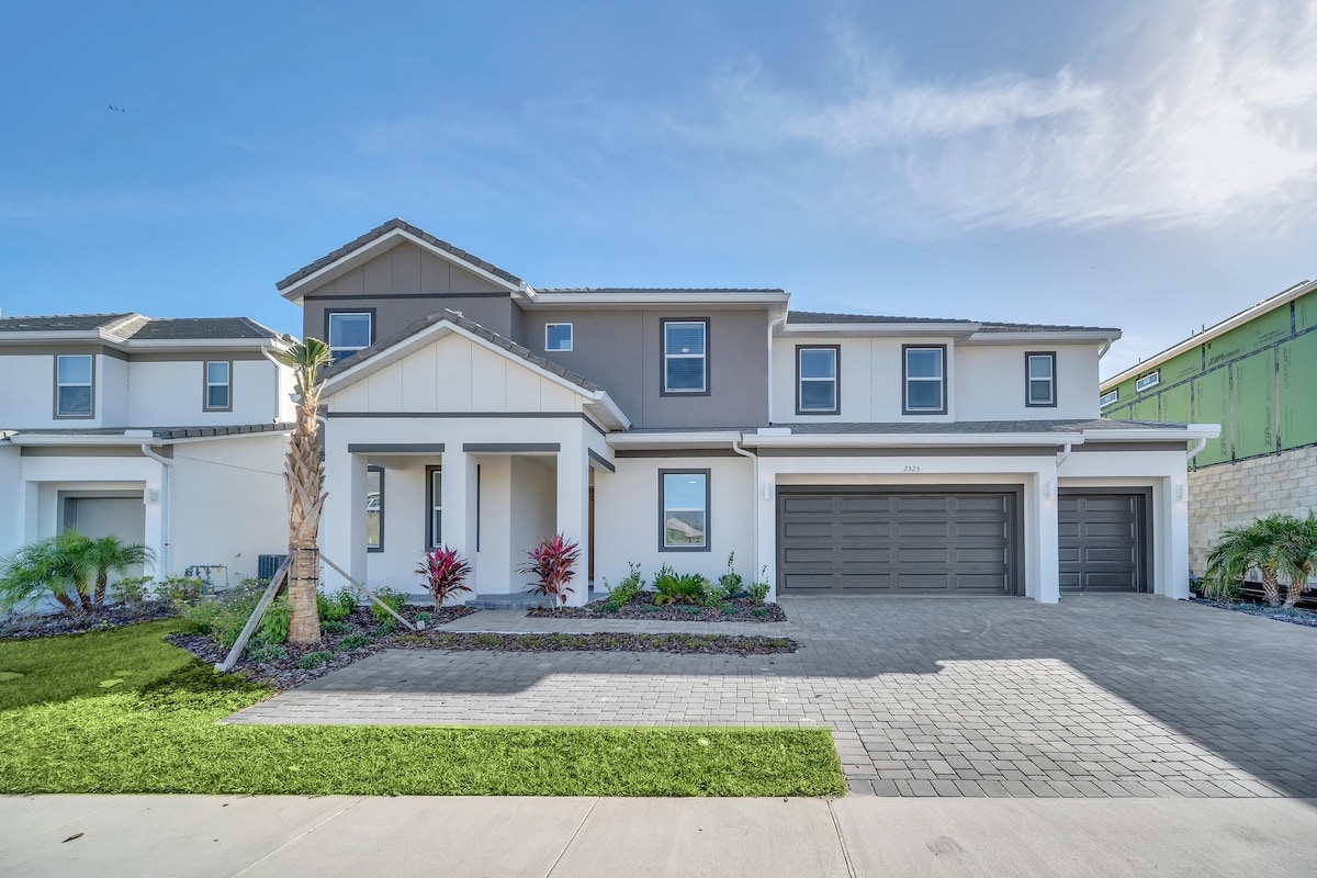 The exterior of a contemporary two-story mansion is displayed, featuring a welcoming front porch, neatly manicured landscaping, and a spacious driveway. Two garage doors are visible, and vibrant plants add a pop of color against the neutral facade, contributing to a modern, clean aesthetic.