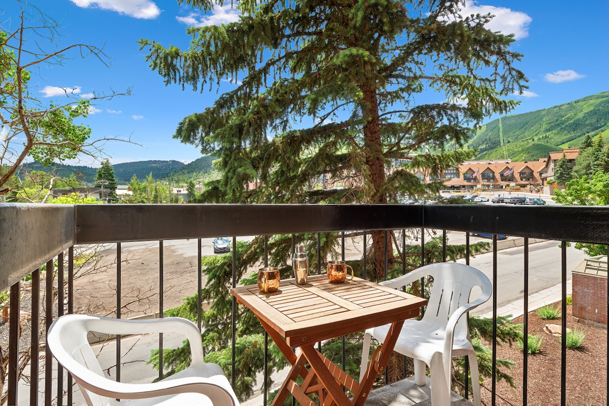 A balcony is showcased with a small wooden table and two white chairs. Green trees and distant mountains create a natural backdrop, while a clear blue sky enhances the outdoor setting. The balcony is enclosed by a black railing.