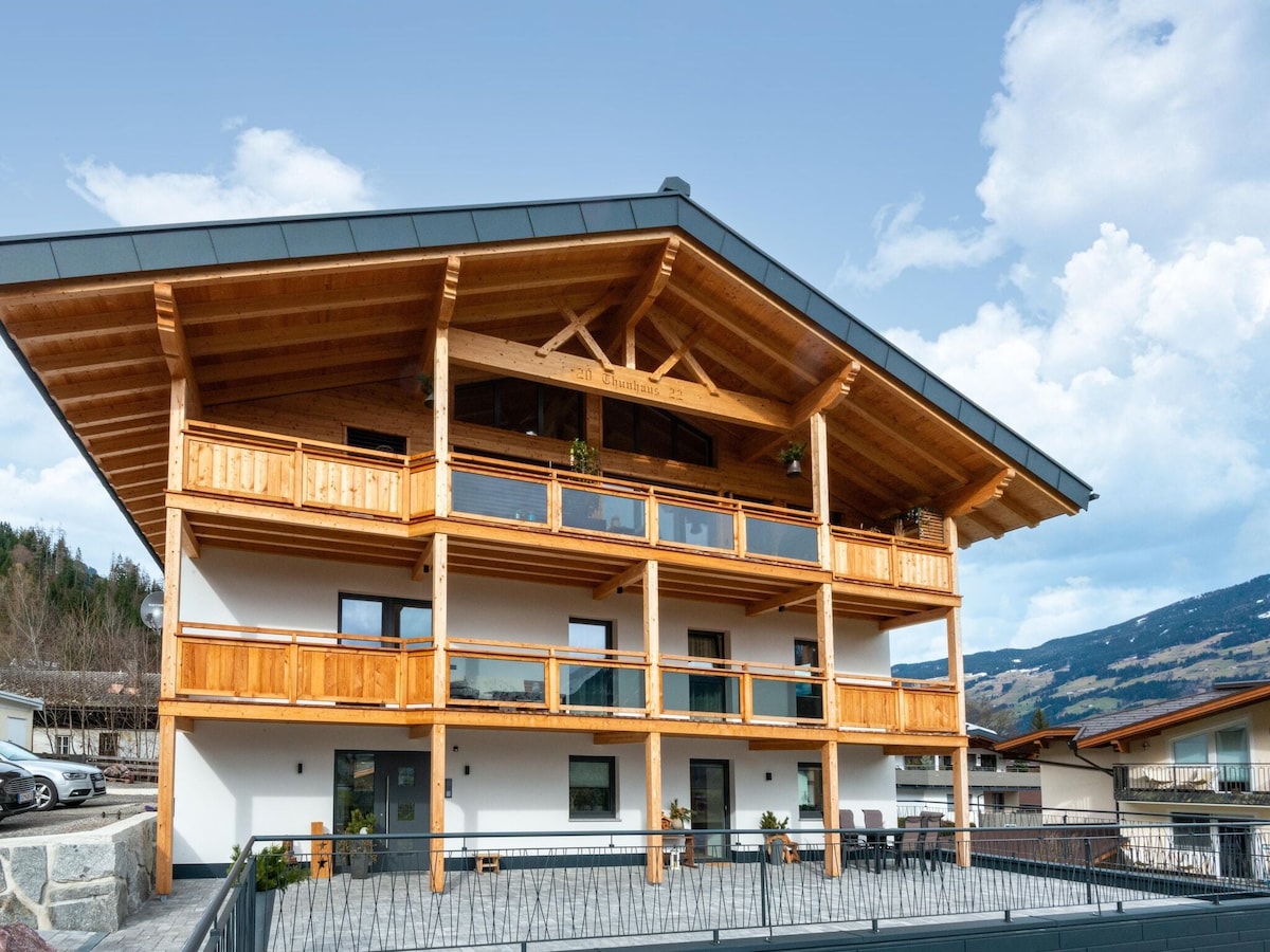 A traditional wooden chalet features multiple balconies with sleek glass railings, showcasing a spacious exterior design. Natural wood accents highlight the structure against a backdrop of mountains under a clear blue sky.