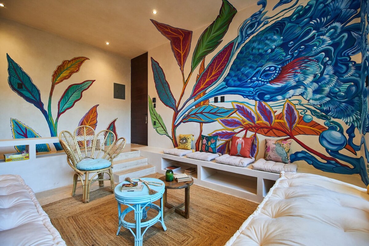 A vibrant living area features artistic wall murals depicting leaves and aquatic life. Comfortable white seating is arranged around a small wooden table, complemented by colorful cushions. Soft lighting illuminates the space, enhancing the natural textures of the furnishings and flooring.