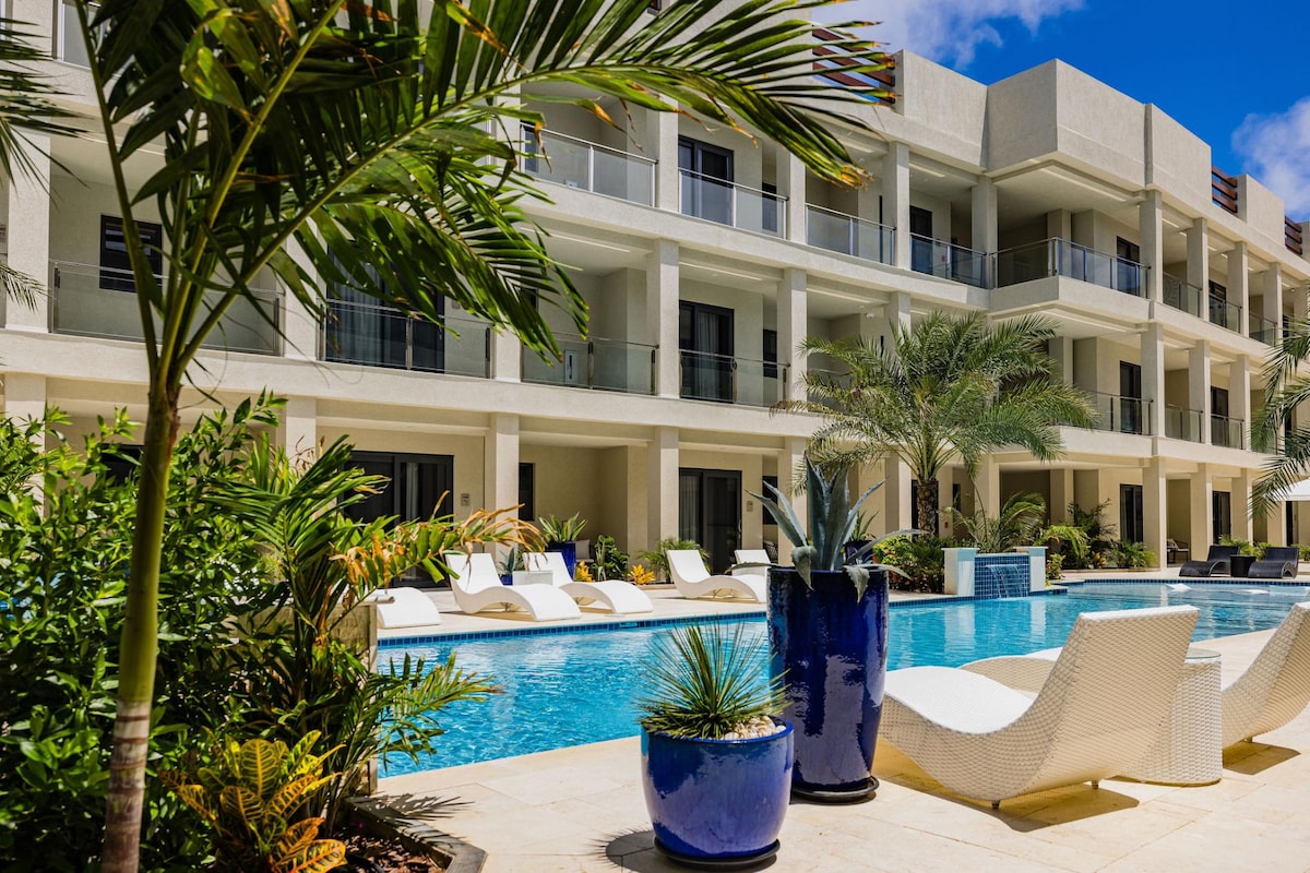 A modern condo complex is featured, showcasing a spacious swimming pool surrounded by lounge chairs and tropical landscaping. The building's light-colored exterior and multiple balconies are visible in the background, while vibrant potted plants add a touch of greenery to the scene.