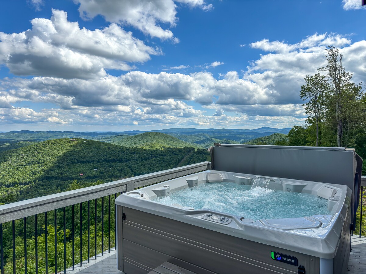 A hot tub is situated on a deck with a railing, offering expansive views of rolling green mountains under a partly cloudy sky. The bubbling water reflects the natural light, creating a serene and inviting outdoor space.