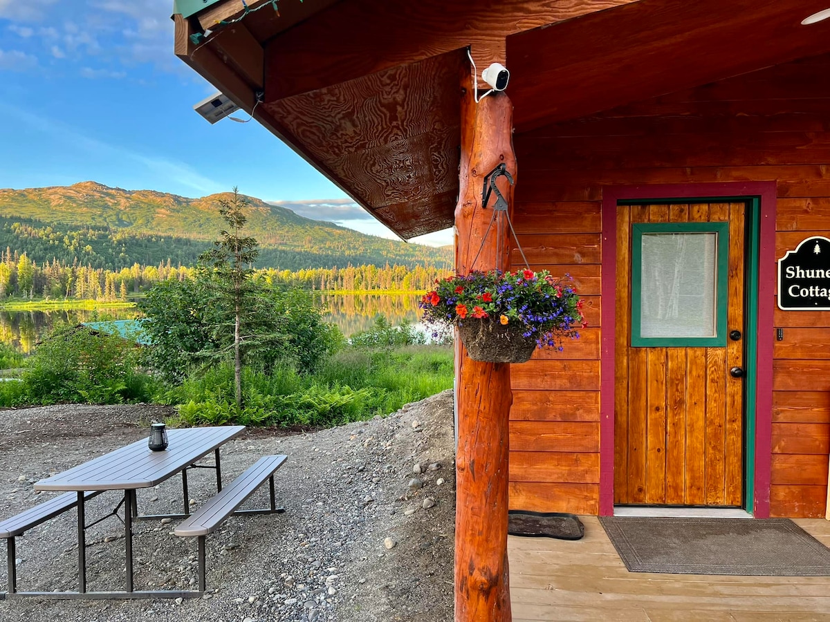 The entrance to Shunem Cottage features a colorful hanging flower basket beside a welcoming entrance door. A picnic table is positioned nearby, overlooking a serene landscape with lake views and lush greenery, framed by distant mountains under a clear blue sky.
