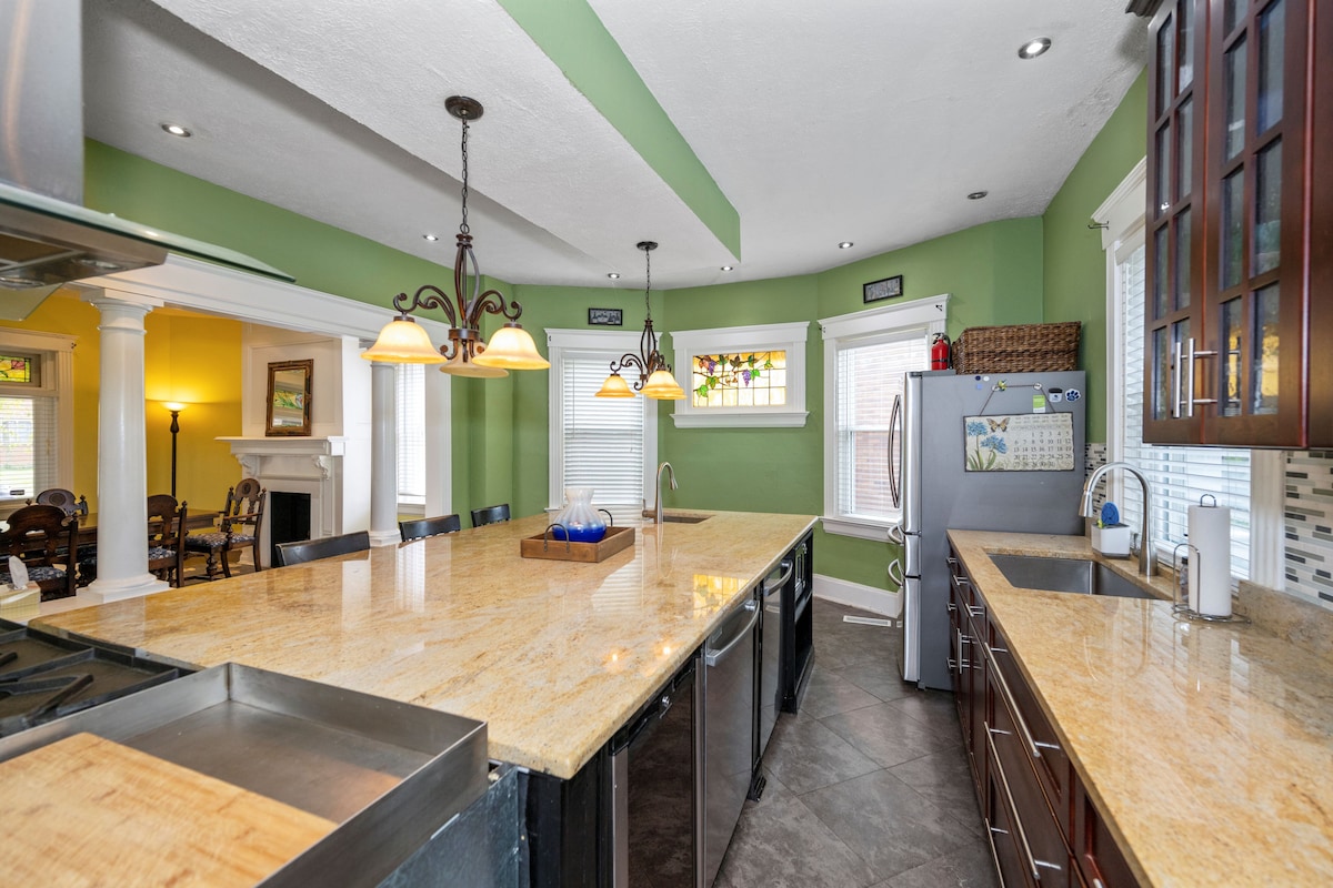 A spacious kitchen features an expansive countertop with a mix of light and dark finishes. Stainless steel appliances are complemented by rich wood cabinetry. Natural light streams in through the windows, highlighting decorative accents over the sink and inviting a warm ambiance.