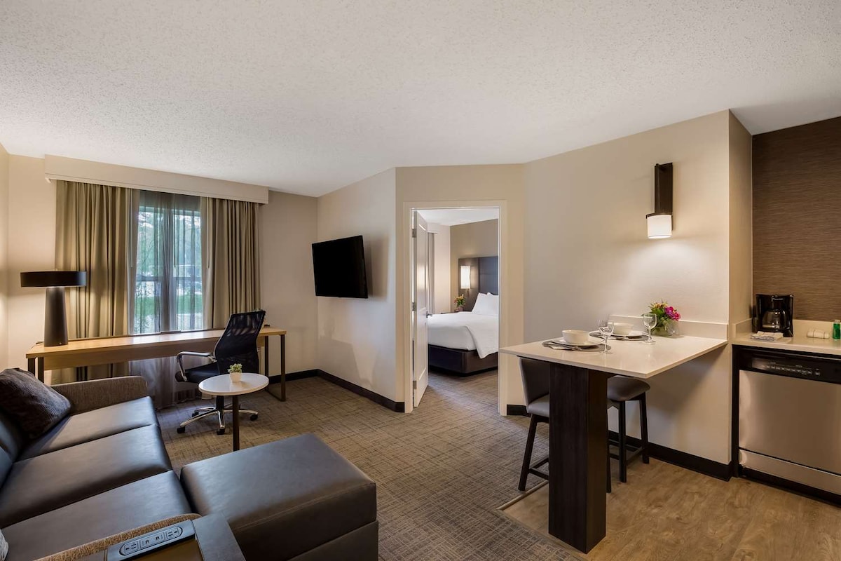 An inviting one-bedroom suite features a living area with a sectional sofa, work desk, and flat-screen TV. A modern kitchen is equipped with a refrigerator, stovetop, and seating area. A separate bedroom is visible through an open doorway, enhancing the spacious layout.