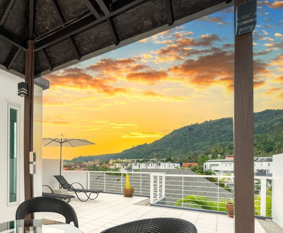 A spacious rooftop terrace is depicted, featuring contemporary outdoor furniture, including a round table with wicker chairs. A sun umbrella provides shade, while lush greenery and sloping hills are visible in the background, under a colorful sunset sky.