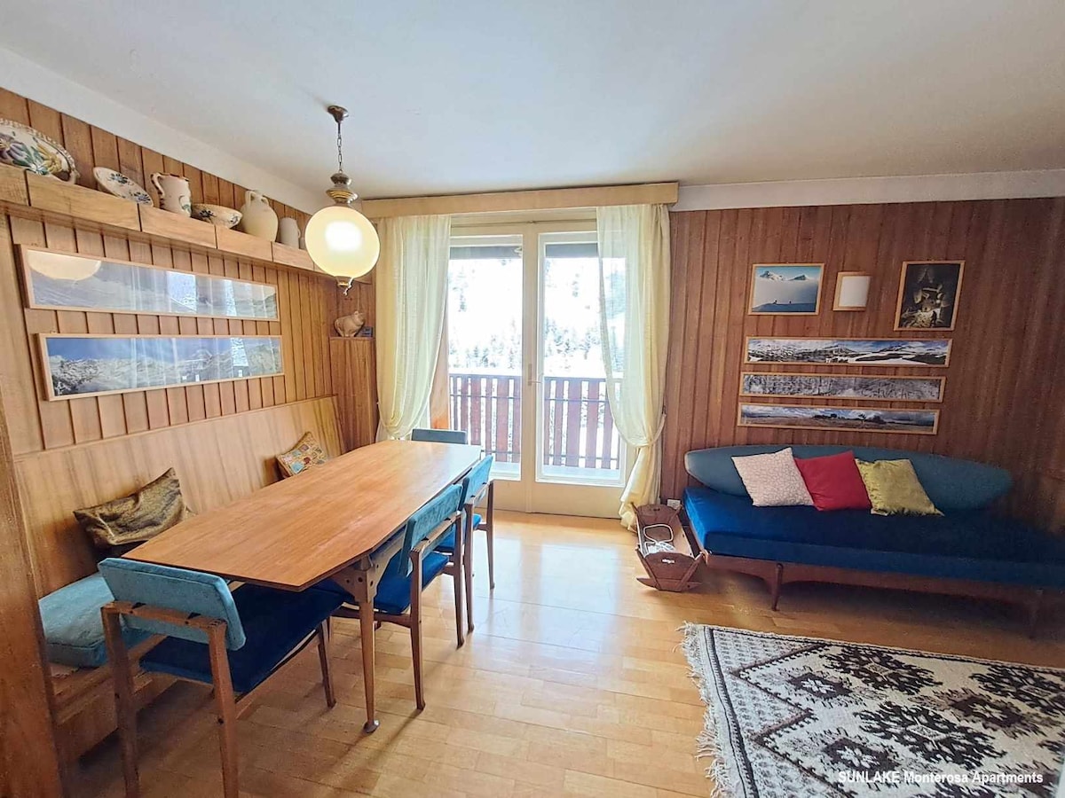 A dining area features a wooden table accompanied by six blue chairs against a backdrop of wood-paneled walls. A large window allows natural light to fill the room, with views of a balcony visible. Decorative wall art adds character to the space.