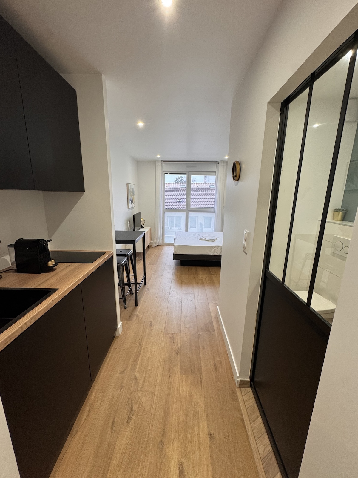 A narrow hallway leads to a bright living space featuring large windows with natural light. On the left, a sleek kitchen area is visible, complemented by minimalist cabinetry. The wooden flooring extends throughout the flat, enhancing the open design.