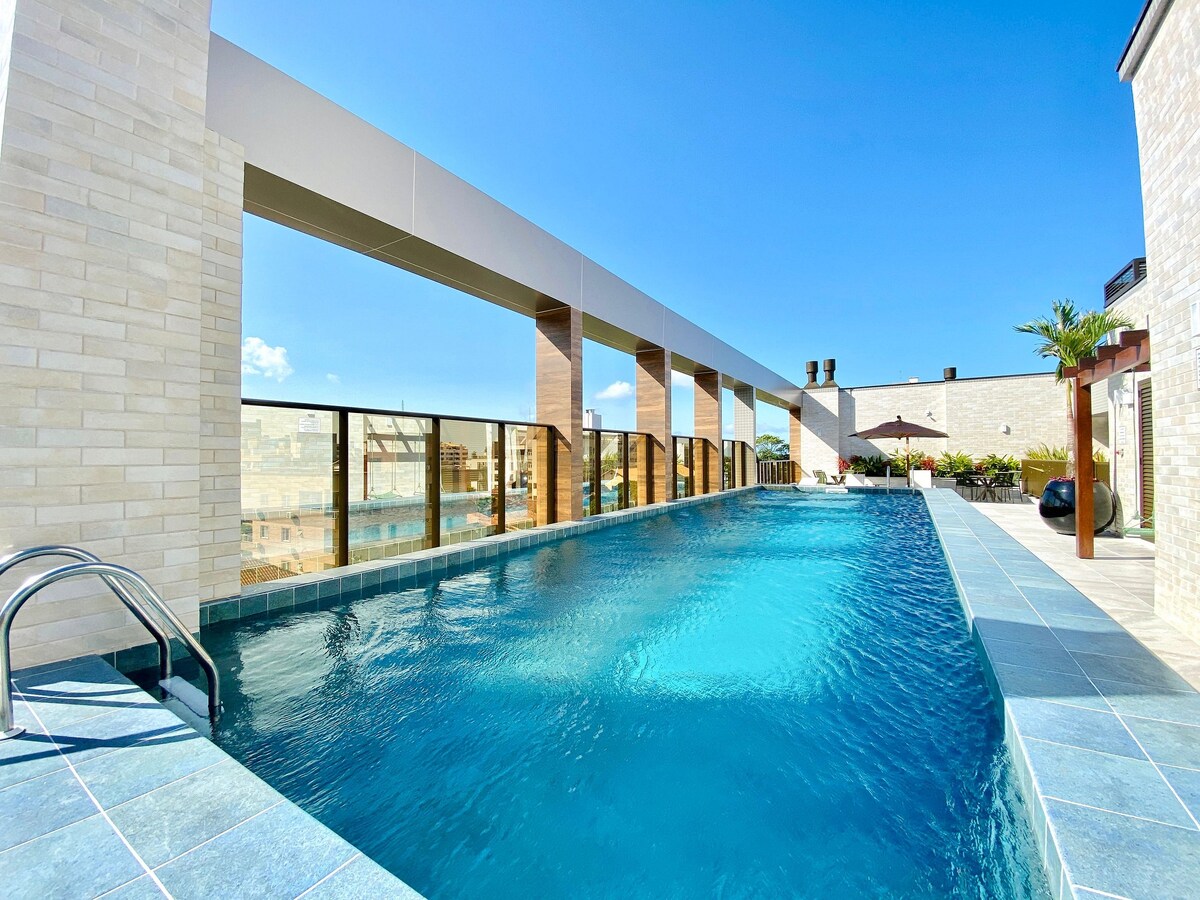 A rooftop pool is seen, surrounded by a glass railing and sunbathing areas. Lounge chairs are positioned alongside the water, with greenery and an umbrella in the background. Clear blue skies reflect on the surface of the inviting pool.