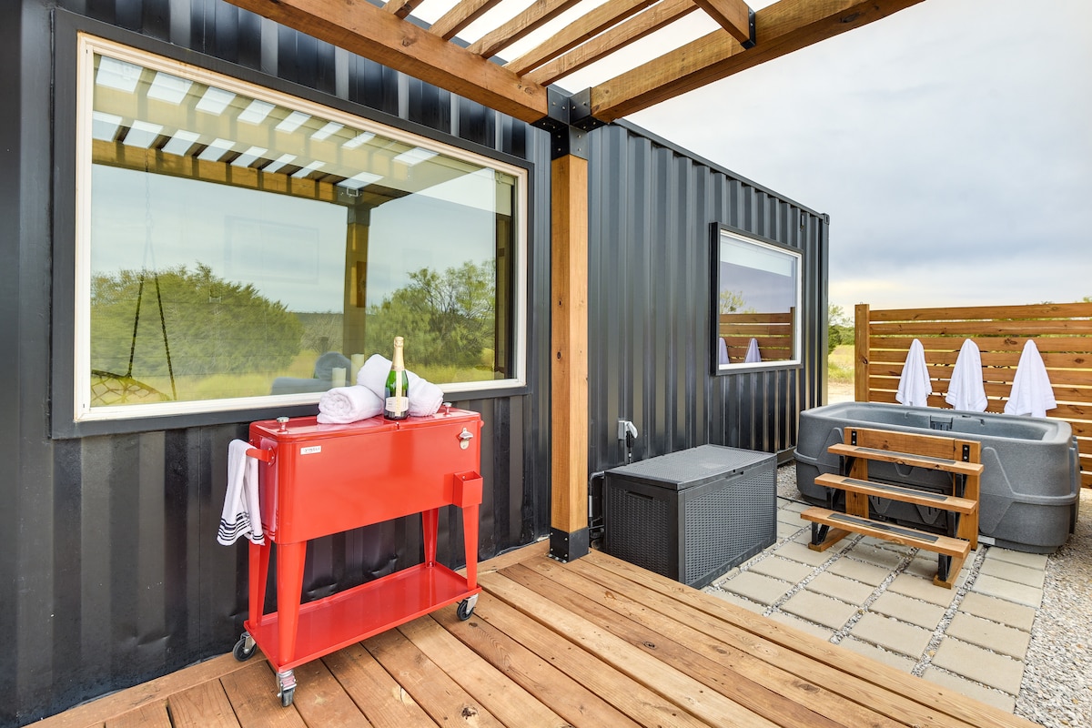 An outdoor deck features a gas grill positioned next to a stylish hot tub. A wooden pergola provides partial shelter, while white lounge chairs enhance the inviting space. The expansive windows of the shipping container reflect the natural surroundings, creating a seamless connection to the landscape.