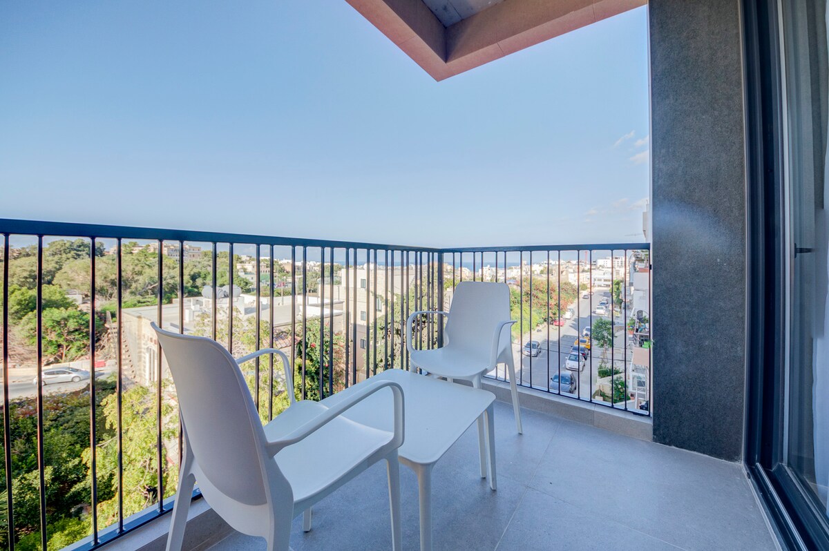 A private balcony features two white chairs and a small table, providing a comfortable spot for relaxation. The railing offers clear views of the serene neighborhood, blending outdoor living with scenic surroundings.