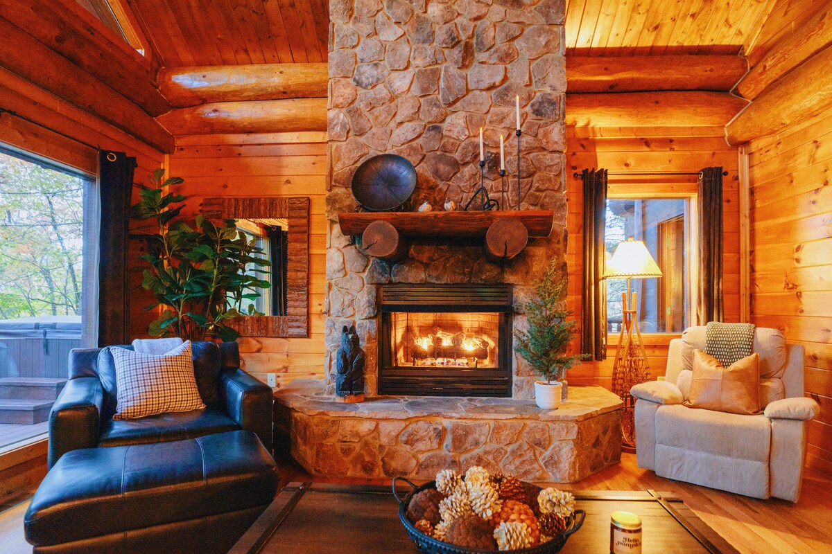 A cozy living area features a stacked stone gas fireplace as its focal point, surrounded by warm wooden walls. Comfortable seating options include a leather sofa and a plush armchair. Natural light streams through large windows, creating an inviting space for relaxation.