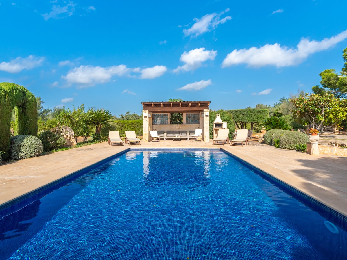 The outdoor area features a large swimming pool surrounded by sun loungers. A pool house is visible in the background, offering shaded seating and a barbecue area. Lush greenery and trees frame the scene, contributing to a tranquil outdoor atmosphere.