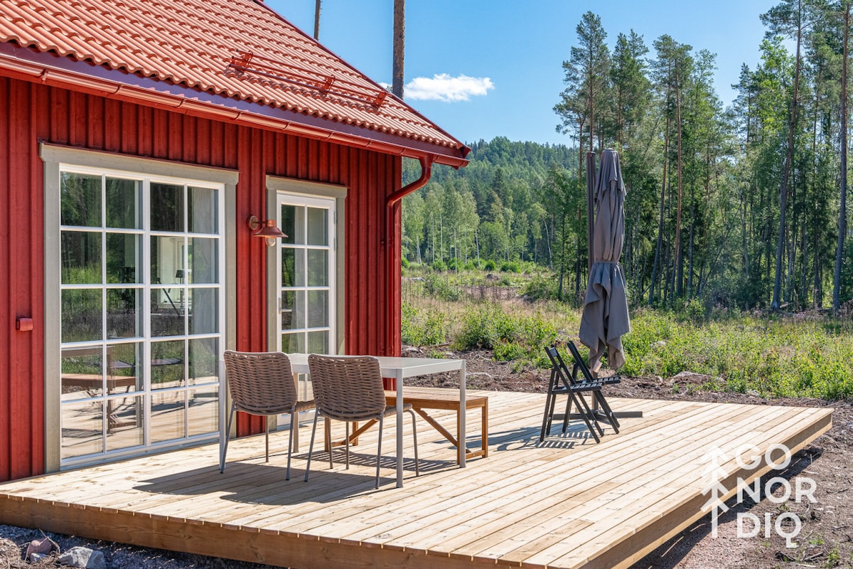 A wooden deck extends from the cabin, featuring a table and two wicker chairs. A folding chair with a protective umbrella is positioned nearby. The backdrop includes lush green forest and a blue sky, enhancing the outdoor living space.
