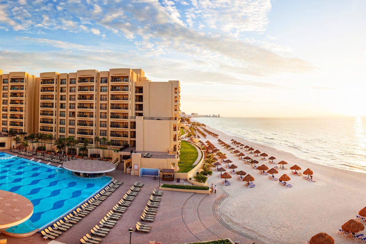An expansive view showcases the resort's large outdoor pool with a wavy design, lined with sun loungers. Beyond, a sandy beach stretches alongside the gentle waves of the ocean, dotted with thatched umbrellas for shade. The warm sunlight casts a golden hue over the scene.