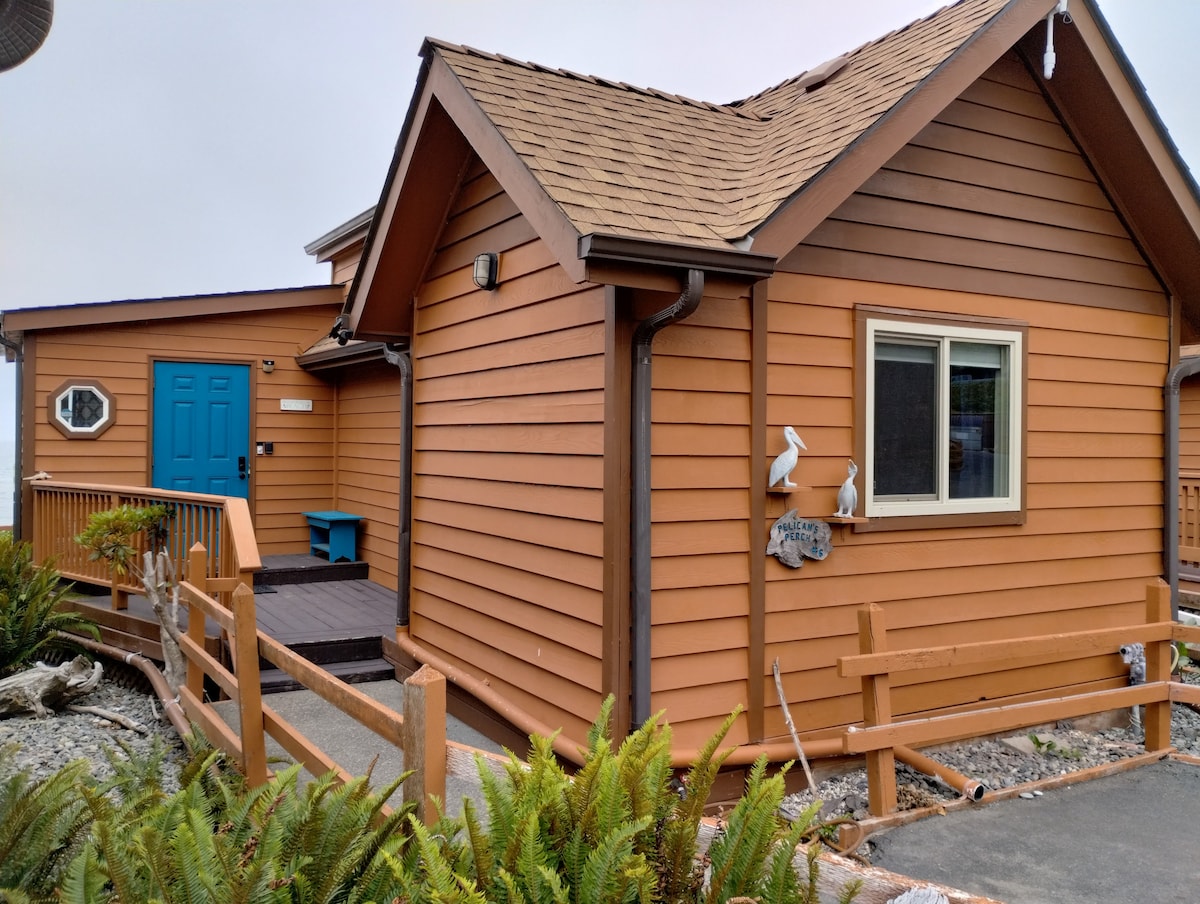 The exterior of the beach cabin is shown, featuring wooden siding in warm earth tones, complemented by a bright blue front door. A small deck with seating is visible, framed by lush greenery and decorative elements such as fish sculptures.