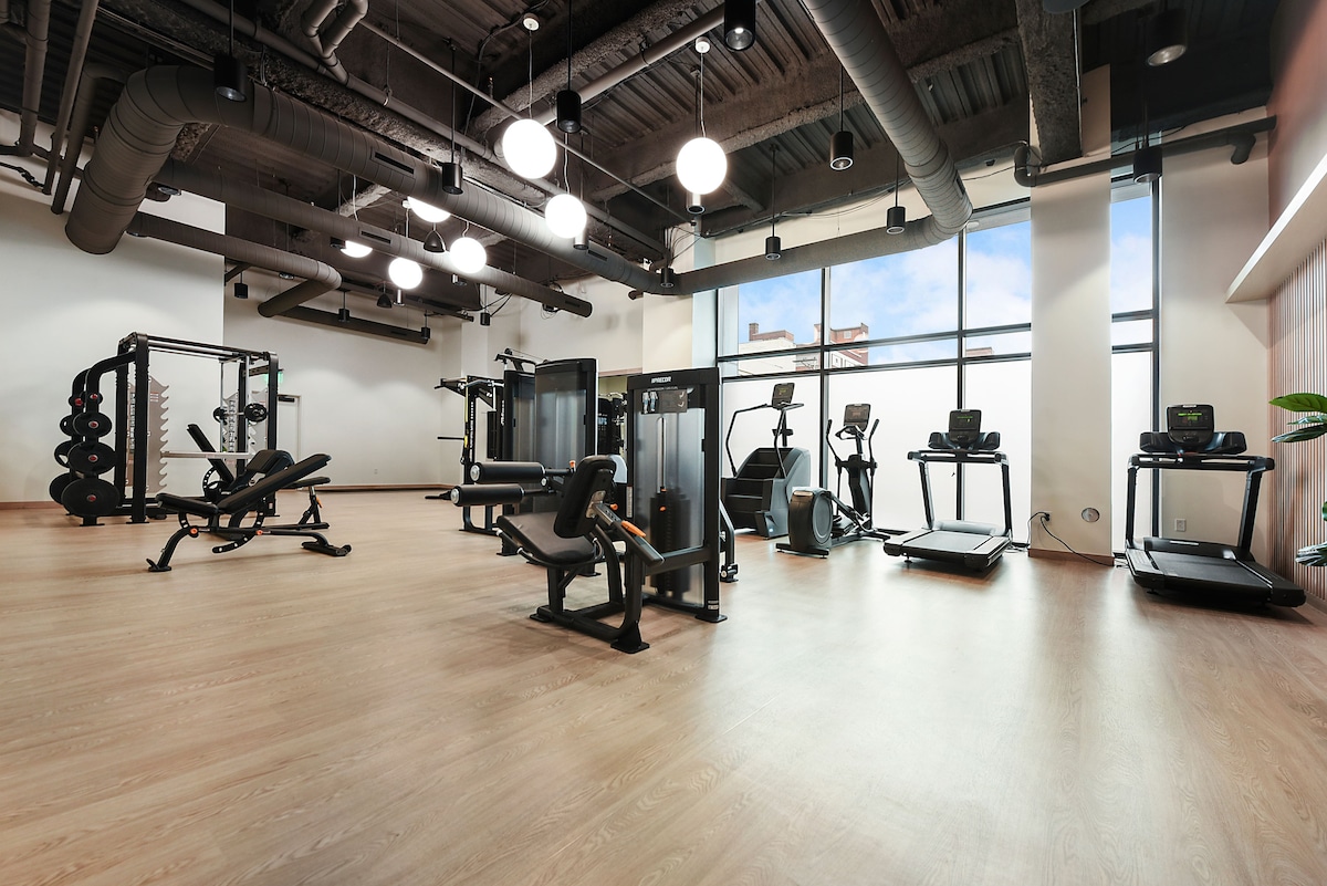 A state-of-the-art fitness center is equipped with cardio machines, strength training equipment, and free weights. Large windows allow natural light to fill the space, creating an open and airy atmosphere. Sleek flooring and modern lighting enhance the contemporary design.
