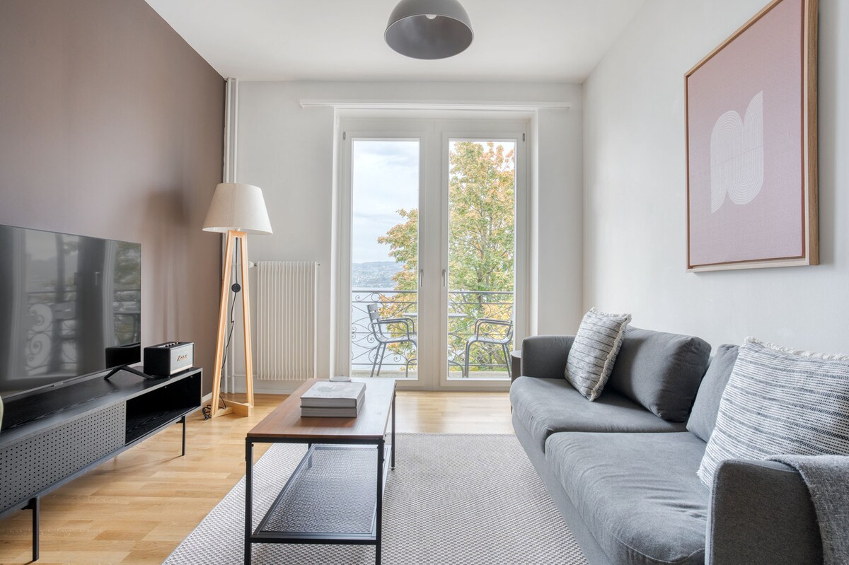 A cozy living area features a comfortable gray sofa, complemented by a wooden coffee table and a flat-screen TV. Large glass doors provide access to a balcony with scenic views, while natural light highlights the warm wood flooring and modern decor.
