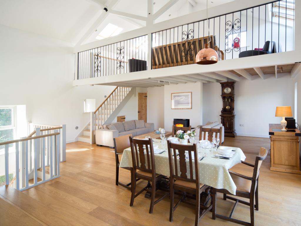 An open-plan living area is highlighted by wooden floors and high ceilings. A dining table set for six is positioned centrally, surrounded by wooden chairs. A comfortable seating area features a large sofa, with a wood burner visible, and a staircase leads to a mezzanine lounge with an ornamental clock.