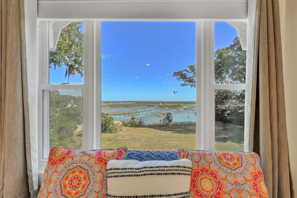 A bright window is framed by soft, textured curtains, offering expansive views of water and greenery. A colorful cushion set against the window adds a cozy touch, inviting relaxation while enjoying the scenic landscape.
