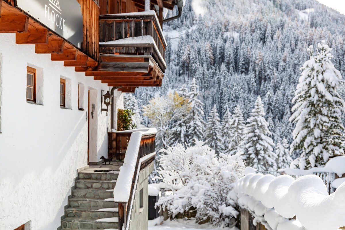 The exterior of the chalet features wooden balconies and a stone staircase leading to the entrance. Snow blankets the surrounding landscape, highlighting the lush pine trees and tranquil atmosphere of the winter scenery.