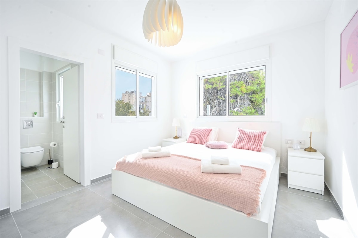 A bright bedroom is presented with large windows offering views of greenery. A spacious bed, dressed in soft linens, is flanked by nightstands with lamps. An ensuite bathroom is visible through a door, showcasing modern fixtures. Neutral tones create a calm atmosphere.