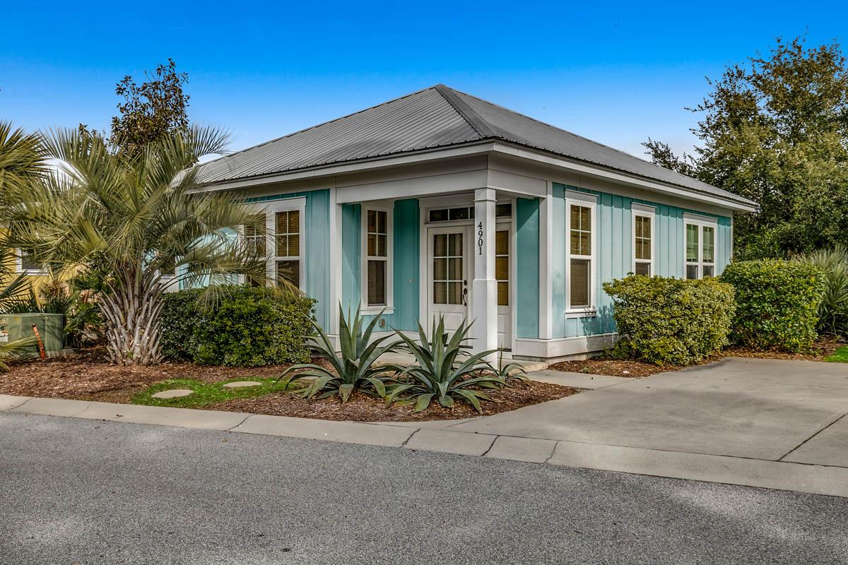 A charming single-story bungalow is shown with a light blue exterior and white trim. Lush greenery surrounds the home, with palm trees and other plants offering a tropical feel. The driveway leads up to the entrance, framed by neatly manicured landscaping.