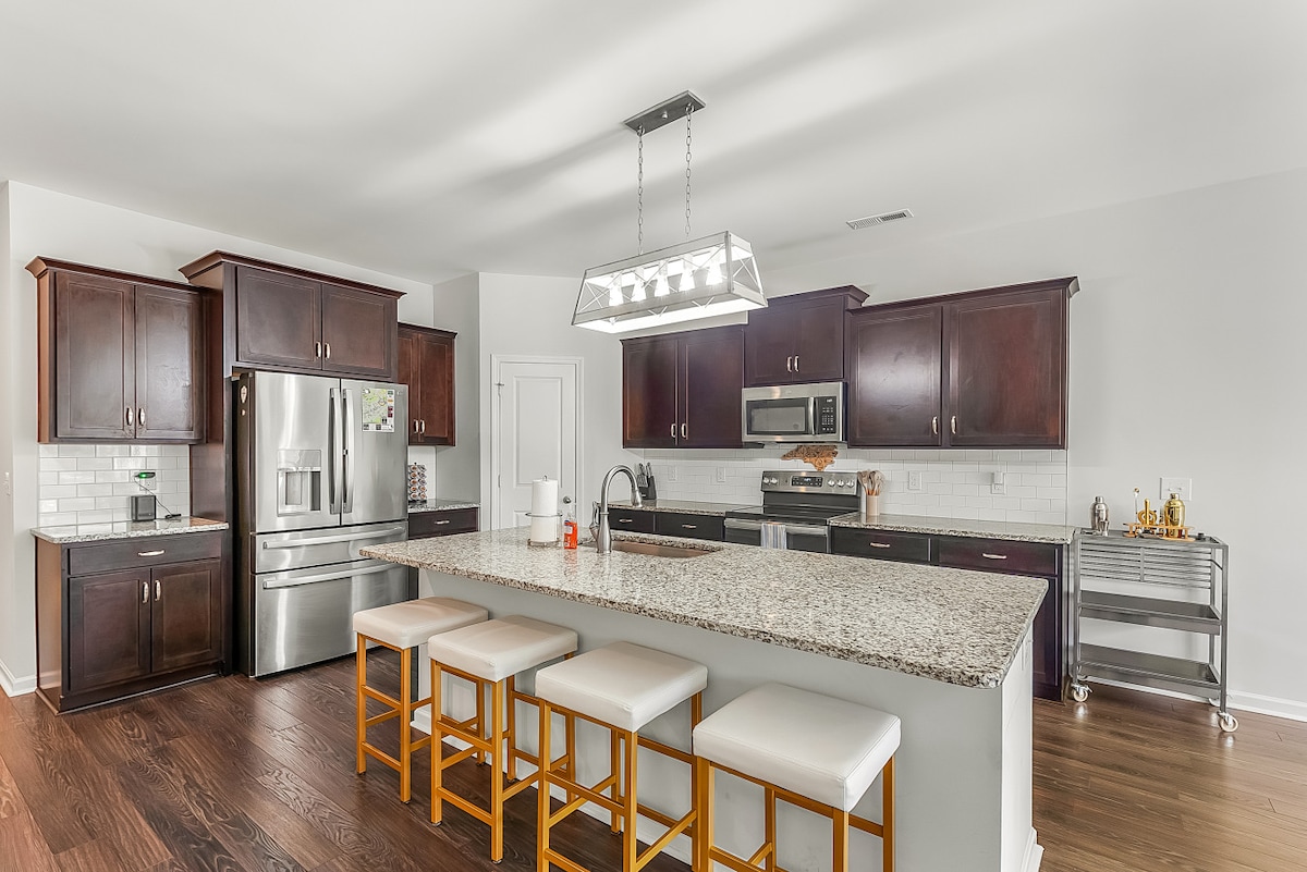 An inviting kitchen features dark wood cabinetry and sleek granite countertops. A spacious island with seating for four is illuminated by a pendant light. Modern appliances, including a stainless steel refrigerator and microwave, are positioned for convenience, while a cart adds extra storage and workspace.
