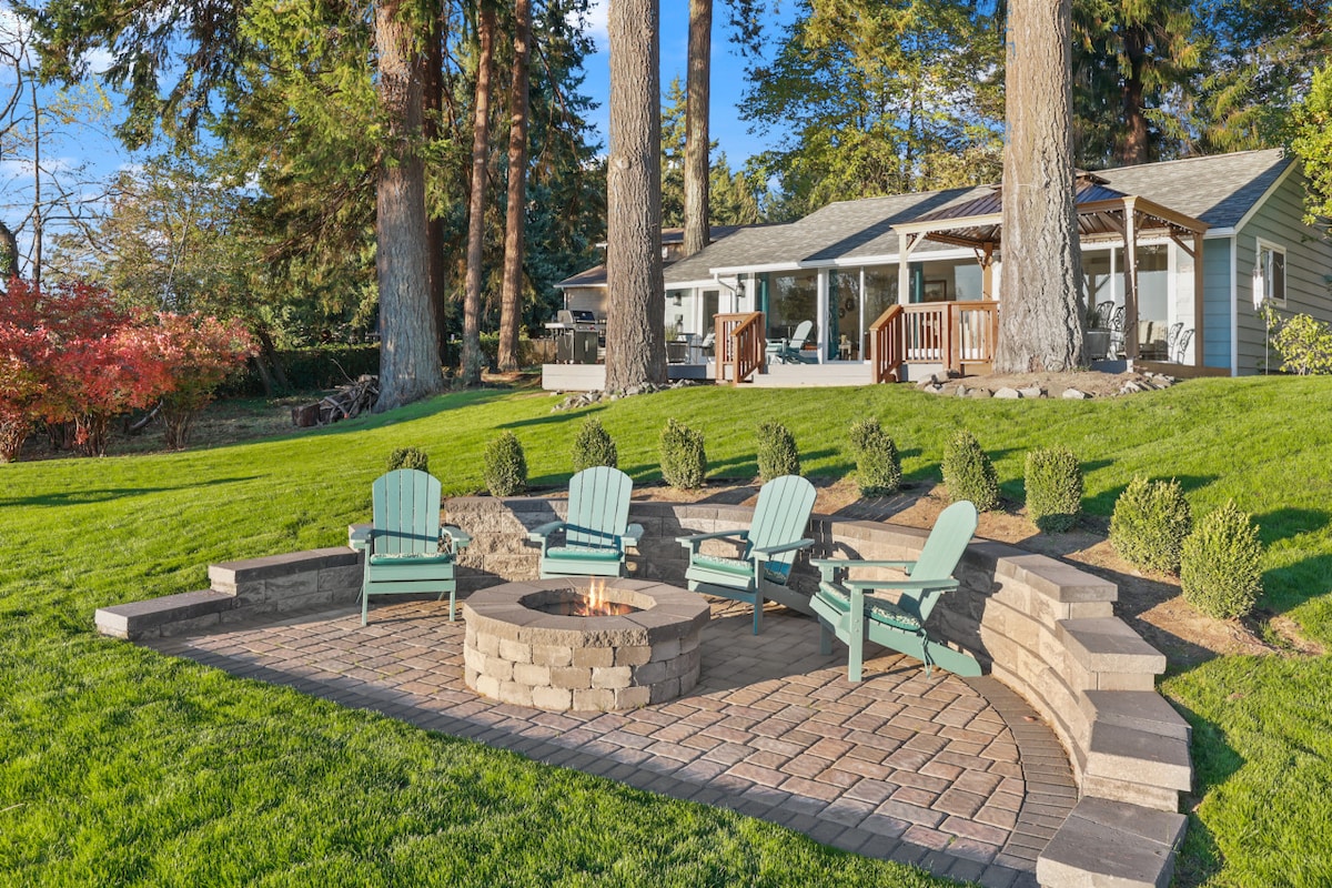 An outdoor seating area is positioned around a stone fire pit, featuring multiple green chairs set on a stone patio. Lush grass and trim hedges outline the space, which is framed by tall trees and a house visible in the background.