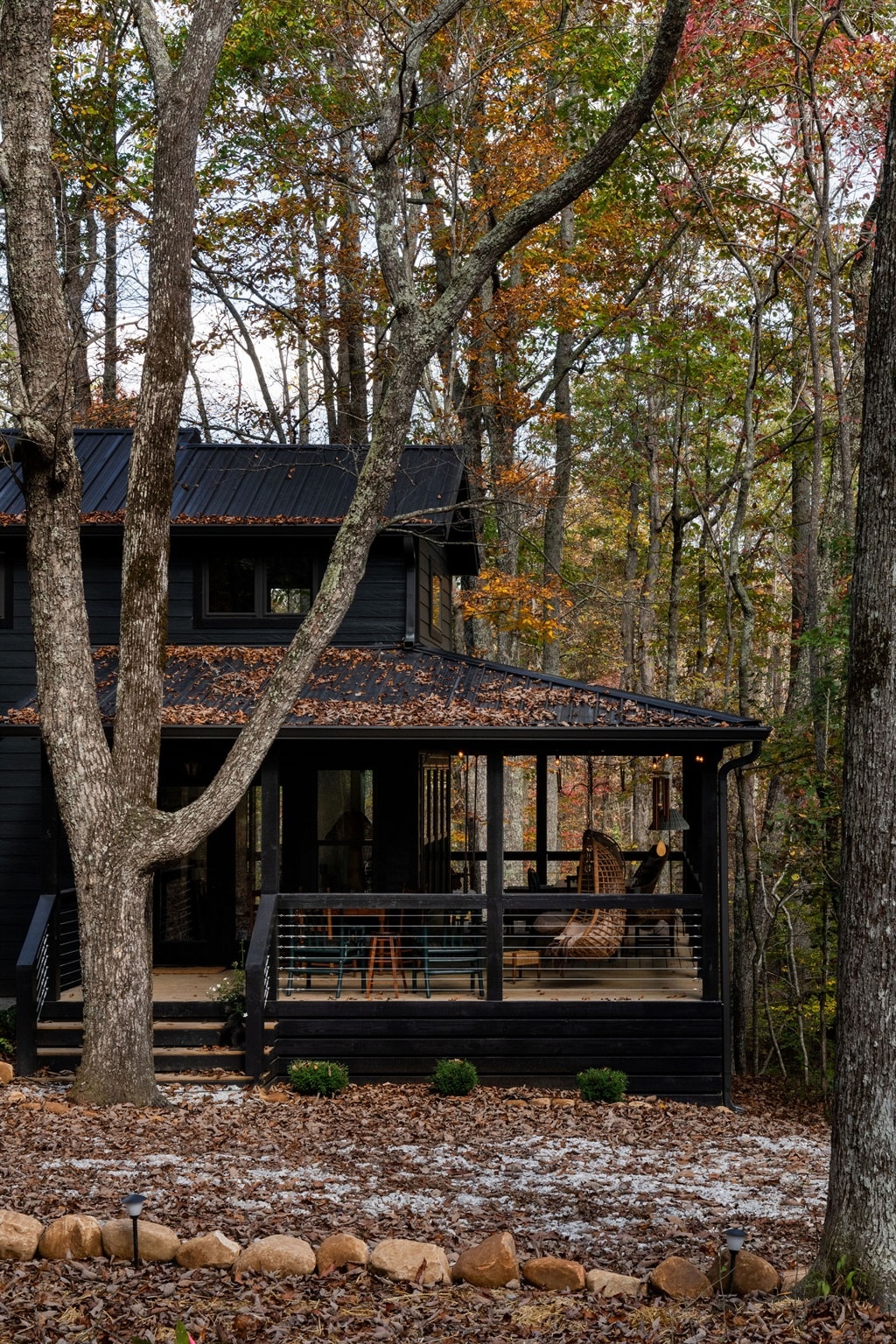 The cabin is nestled among tall trees adorned with autumn foliage, displaying a mixture of warm hues. A spacious covered porch features a swing and offers views of the surrounding nature. Leaf-strewn ground leads to the entrance, enhancing the serene, rustic atmosphere.