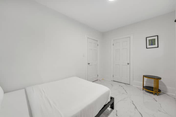 Dhomë gjumi private minimaliste me një krevat elegant me madhësi të plotë, ambiente të brendshme të bardha të ndritshme dhe një tavolinë anësore moderne, e përsosur për një qëndrim të rehatshëm dhe çlodhës.