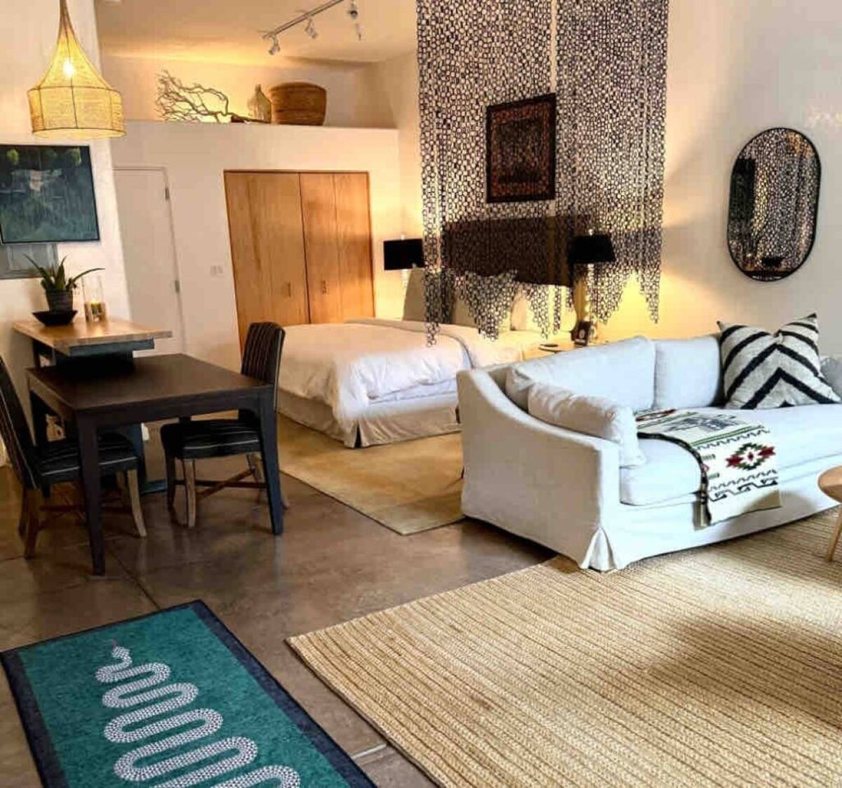 A spacious studio layout is highlighted by a comfortable seating area featuring a light-colored sofa and a dining table for four. The sleeping area is separated by decorative hanging elements. Soft lighting and natural textures create a relaxed environment, complemented by earthy hues in the decor.