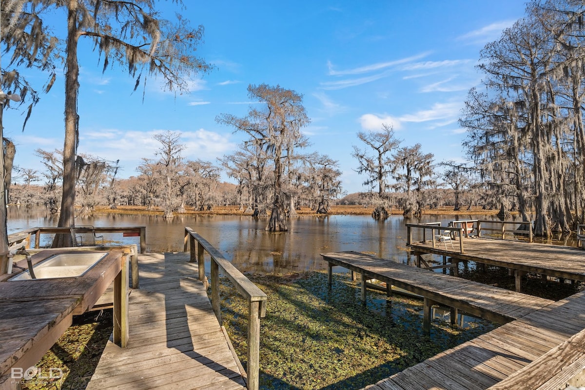 A wooden boardwalk leads to a serene lake surrounded by cypress trees draped in moss. The calm water reflects the sky, while a few boat docks are visible, providing access to the lake for guests. Natural foliage adorns the scene, enhancing its tranquil appeal.
