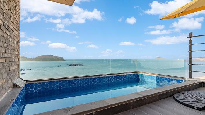 A small blue-tiled pool is situated on a terrace, offering expansive views of the ocean. The glass railing allows unobstructed sightlines to the water, framed by a clear sky with scattered clouds. Sunshades provide shelter on sunny days.