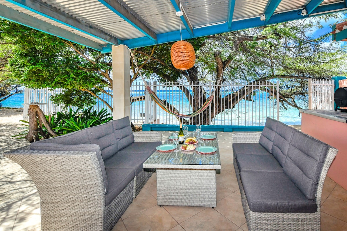 A shaded outdoor seating area features a grey woven sectional sofa surrounding a glass-topped table. The space is complemented by a hammock nearby, with views of the turquoise sea visible through the surrounding foliage.
