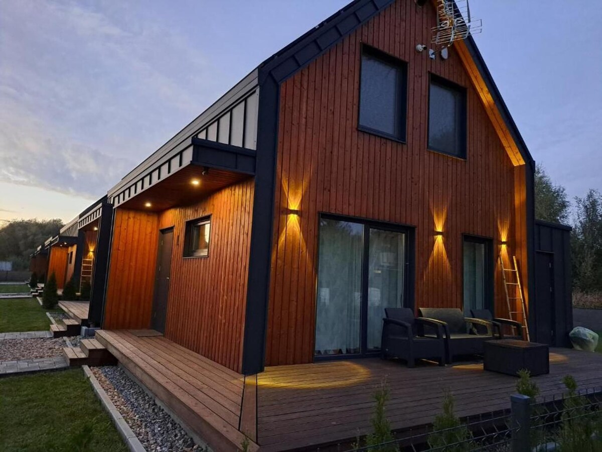 A modern semi-detached house features a wooden exterior and large glass doors. Soft lighting highlights the entrance and porch area. The deck is furnished with comfortable seating, providing a welcoming spot for relaxation. A landscaped garden surrounds the property, enhancing the outdoor space.
