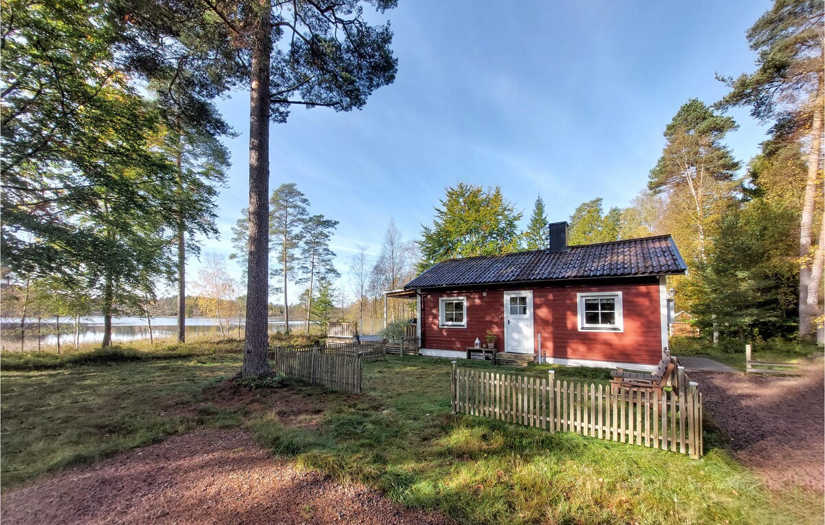 A red vacation home is surrounded by greenery, featuring large windows on one side and a chimney. A wooden fence encloses a small patio, while the serene Lake Hulta is visible in the background through the trees.