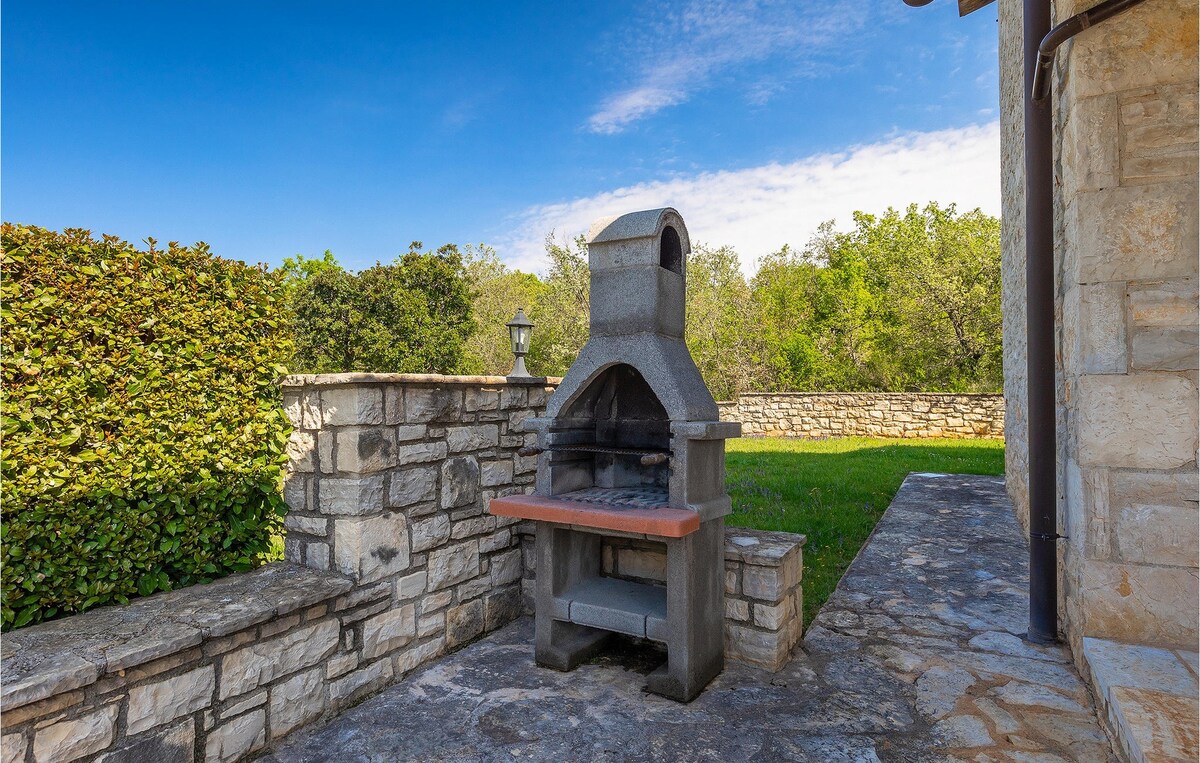 An outdoor barbecue grill is situated on a stone patio, surrounded by greenery and lawn. The grill features a brick top and a hood, providing a functional space for outdoor cooking. A wall of smooth stone borders the area, enhancing the rustic charm.